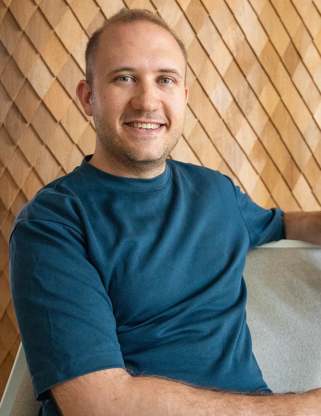Ein junger Mann mit kurzem Haar und einem blauen T-Shirt sitzt entspannt auf einem Sofa vor einer holzverkleideten Wand und lächelt.