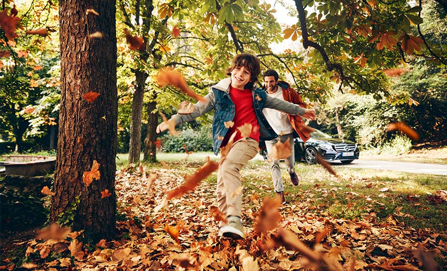 Kind spielt mit seinem Vater im Blätterhaufen im Hintergrund ein Mercedes-Benz
