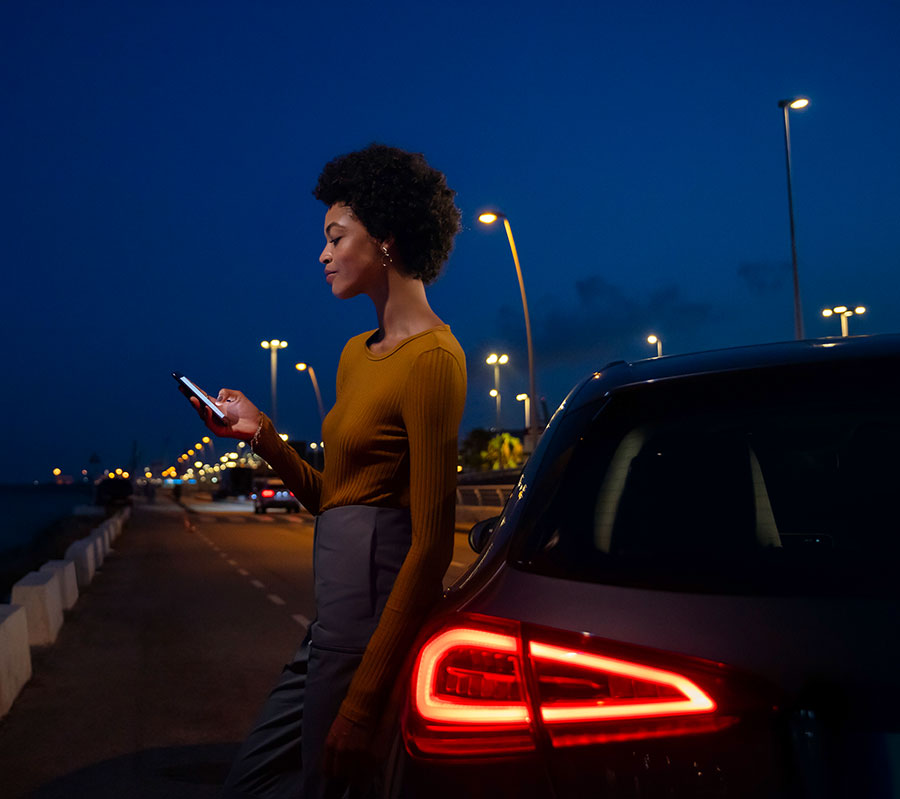 Frau lehnt lässig mit einem Handy in der Hand an ihrem Mercedes-Benz auf einer nächtlichen Straße