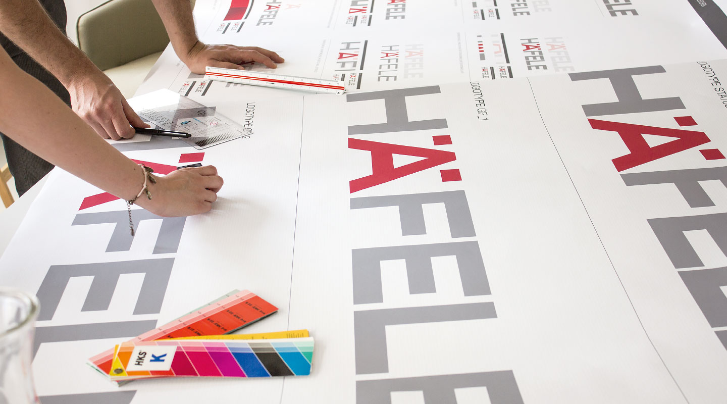 Designer prüfen ausgedruckte Häfele Schriftzüge mit Lineal und HKS Farbfächer.