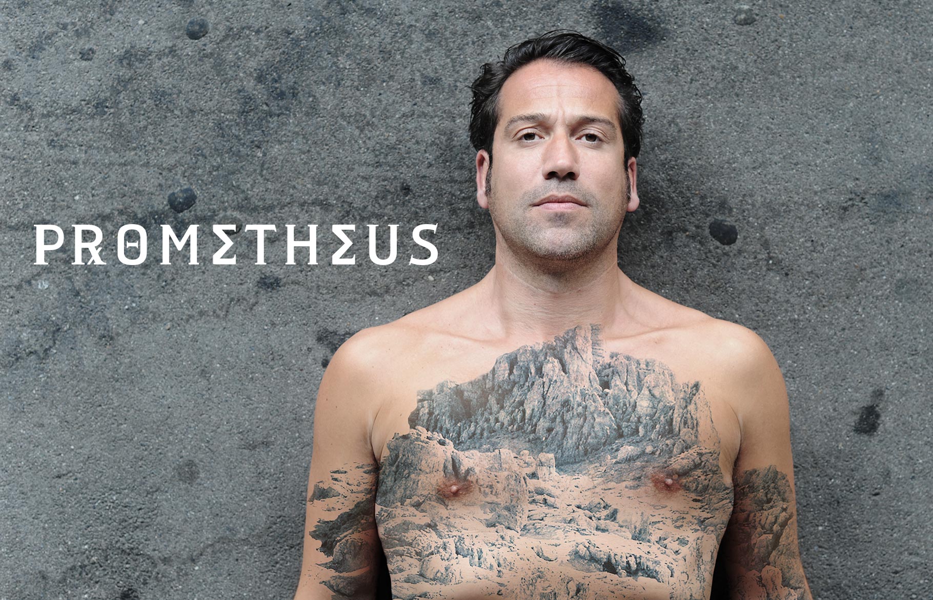 Foto für 'Prometheus': Ein Mann liegt auf dem Asphalt. Auf seinem nackten Oberkörper ist ein großflächiges Tattoo einer Landschaft zu sehen.